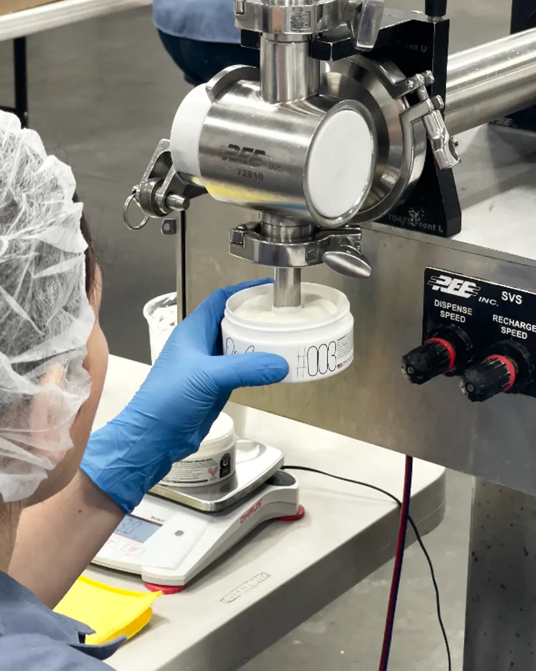 Gloved hand holding a #003 jar under the filling machine, above a precision scale